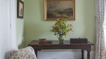 Una habitación con una pared verde, un cuadro de paisaje enmarcado, un sillón floral, una consola de madera con un florero y un sillón verde.