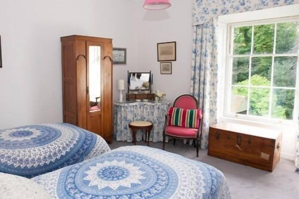 A bedroom with two beds, a wooden wardrobe, a small table, a chair, and a window with a view of greenery.