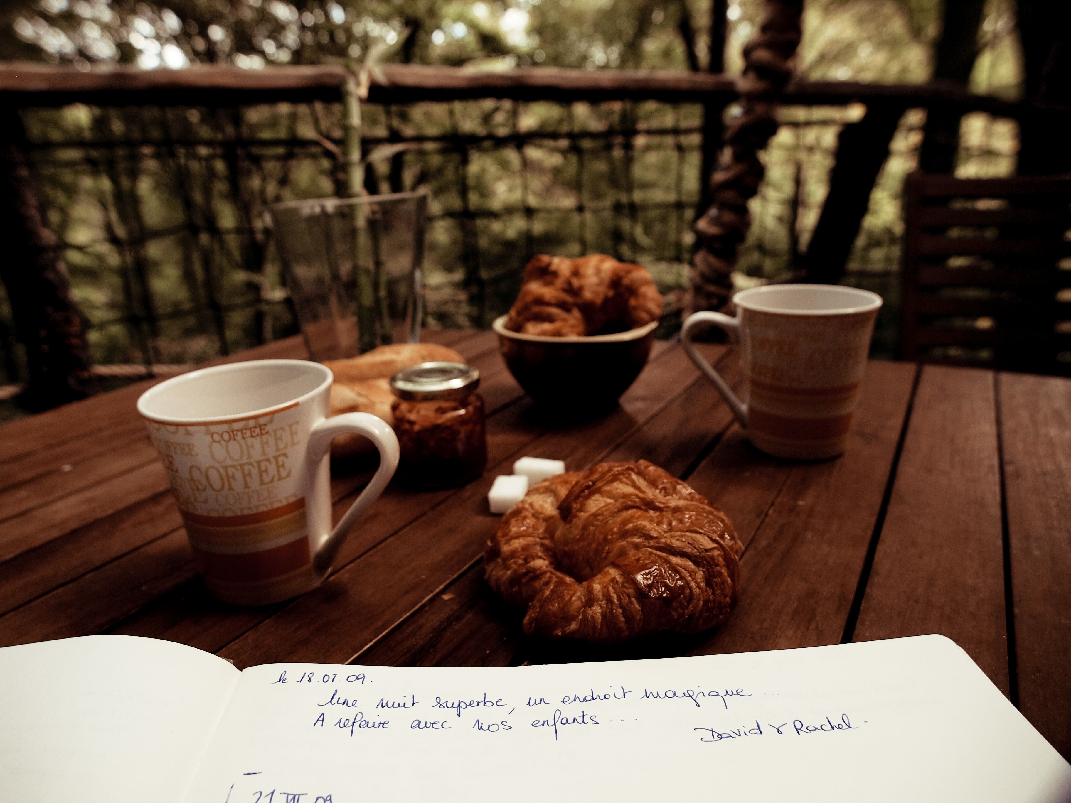 Ein Holztisch mit einer Kaffeetasse, einem Croissant, einer Marmeladenglas und einer Schüssel Kekse.