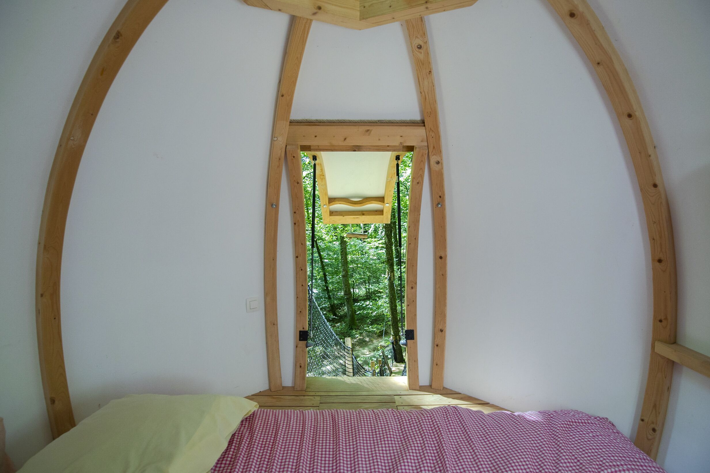 A bedroom with a wooden ceiling, a bed with a checkered bedspread, and a window with a view of a forest.