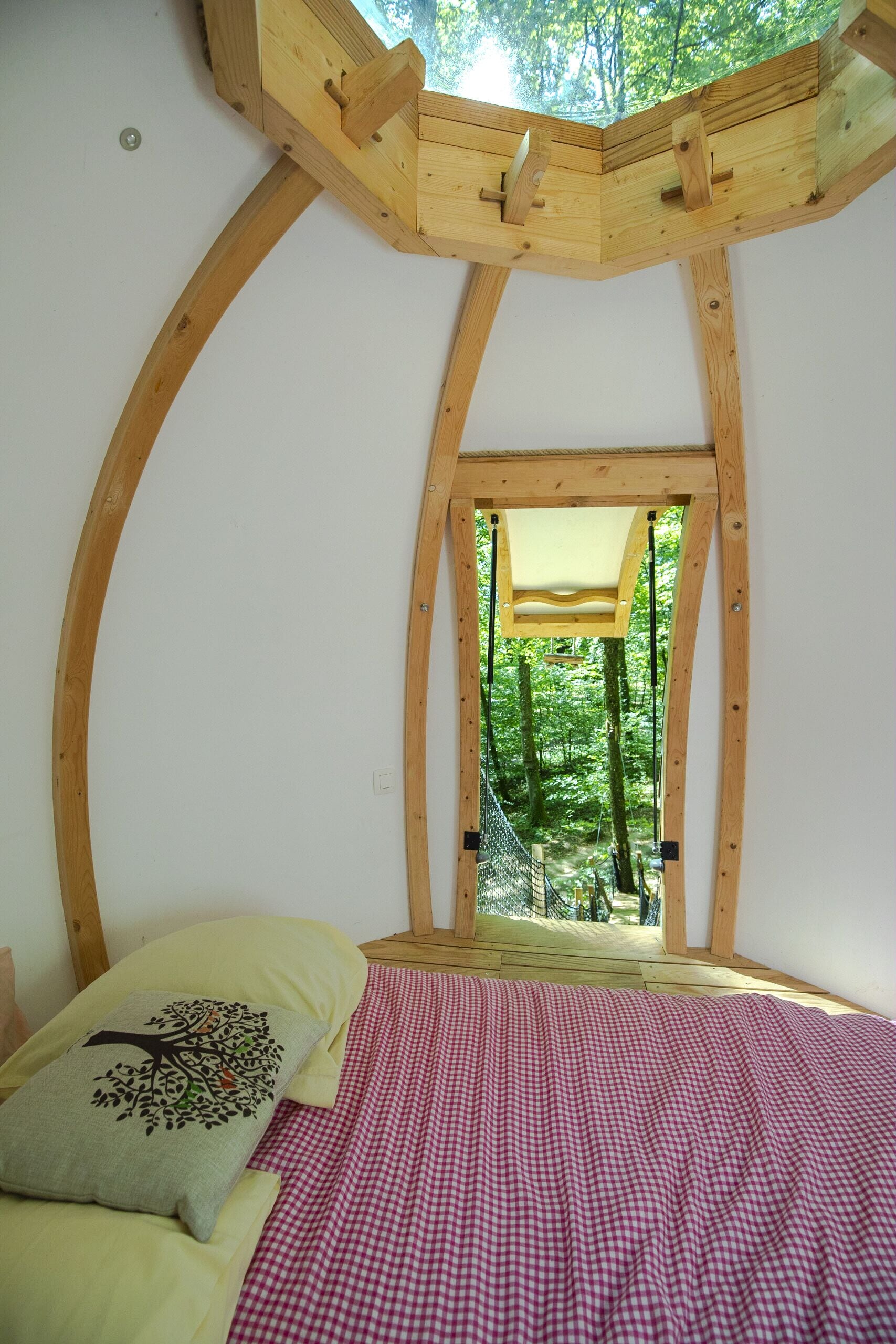 Ein Schlafzimmer mit kariertem Bettbezug, einem Kissen mit Baummotiv und Blick auf einen Wald durch einen Holzrahmen.