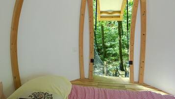 Ein Schlafzimmer mit kariertem Bettbezug, einem Kissen mit Baummotiv und Blick auf einen Wald durch einen Holzrahmen.