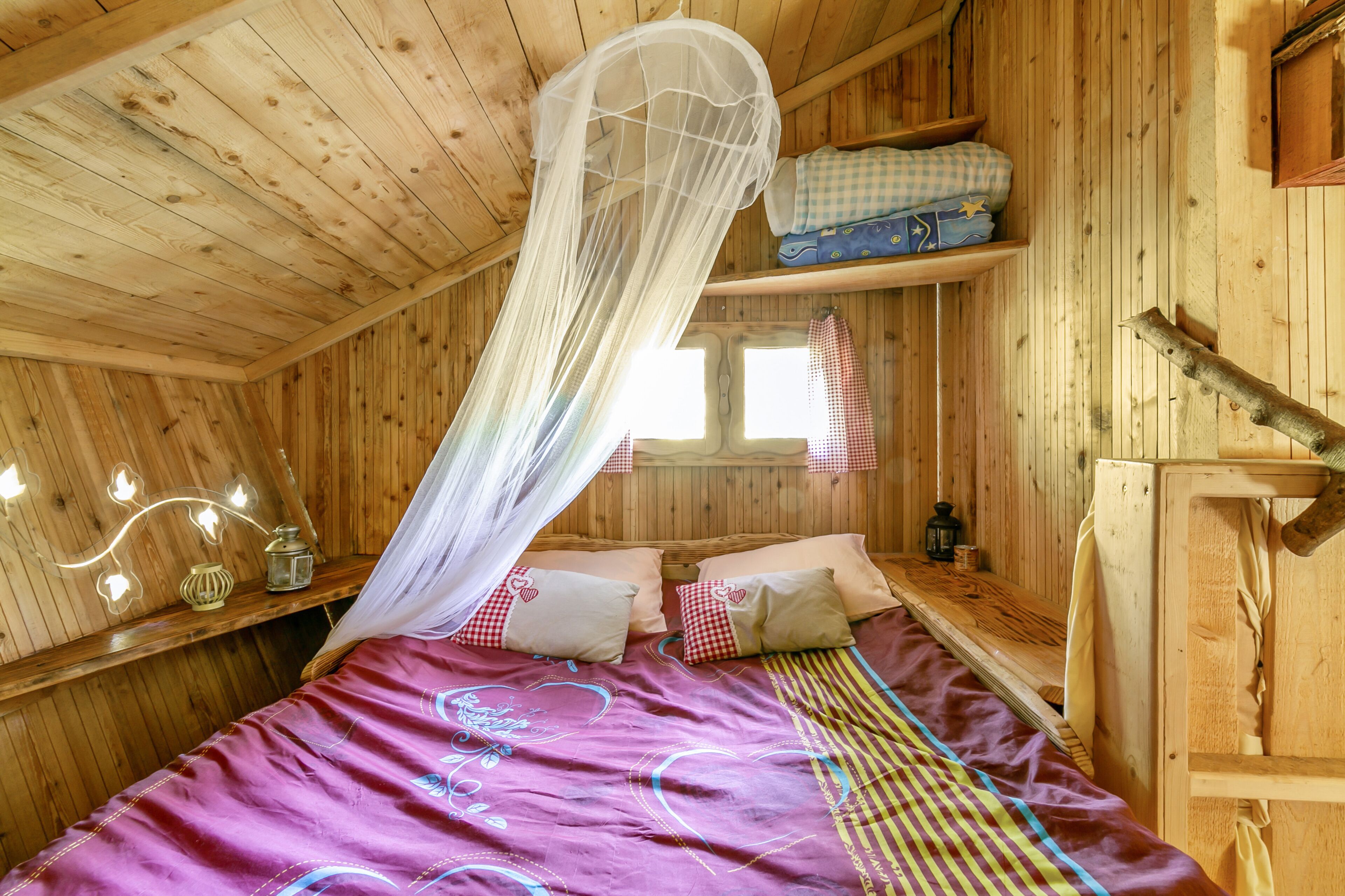 Un dormitorio de cabaña de madera con una cama, un dosel, un estante con ropa doblada y una ventana.