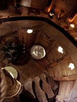 Una habitación circular y acogedora de madera, con una zona de estar central con cojines, vigas de madera y una pequeña mesa con una vela.