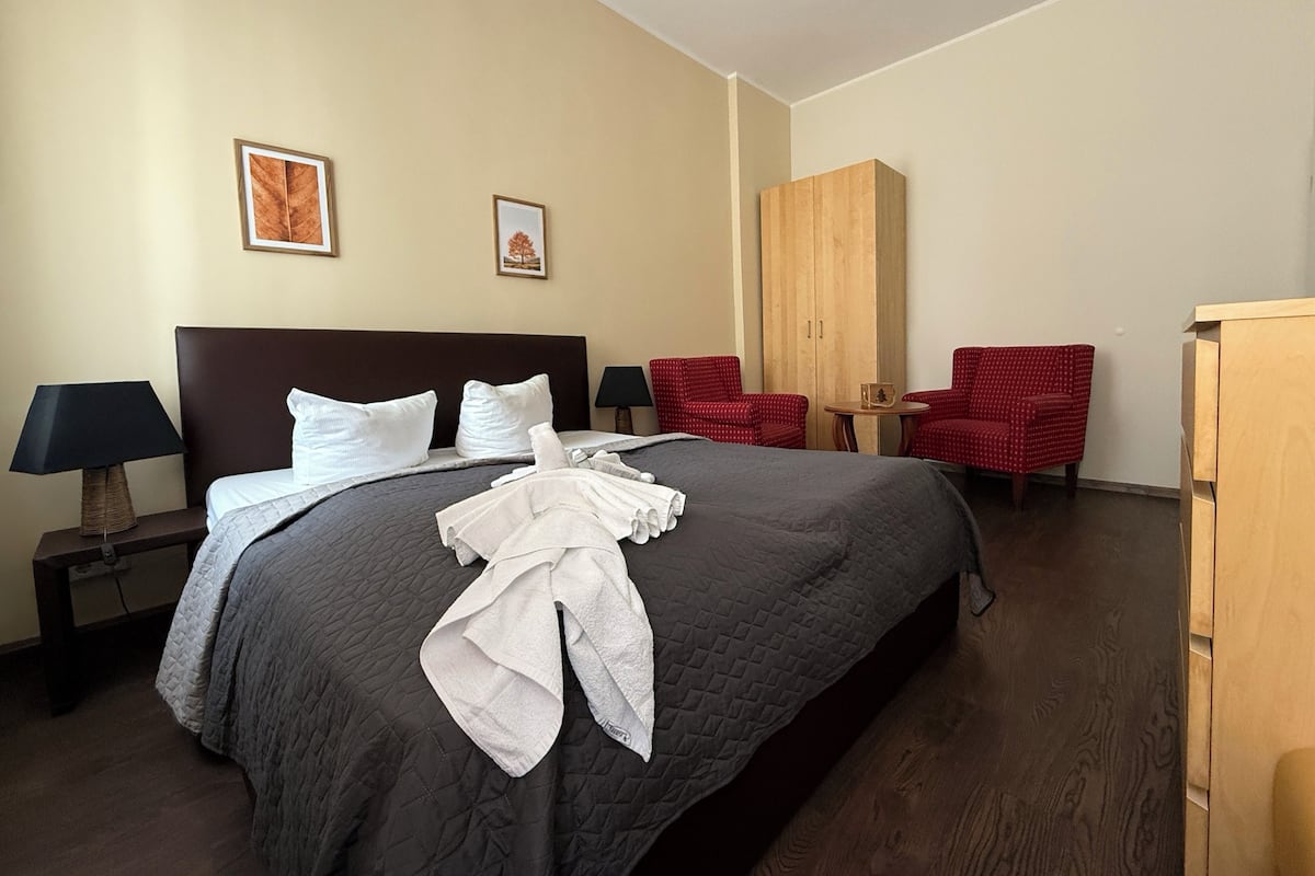 Habitación de hotel con una cama grande, dos sillas rojas, un armario de madera y dos cuadros en la pared.