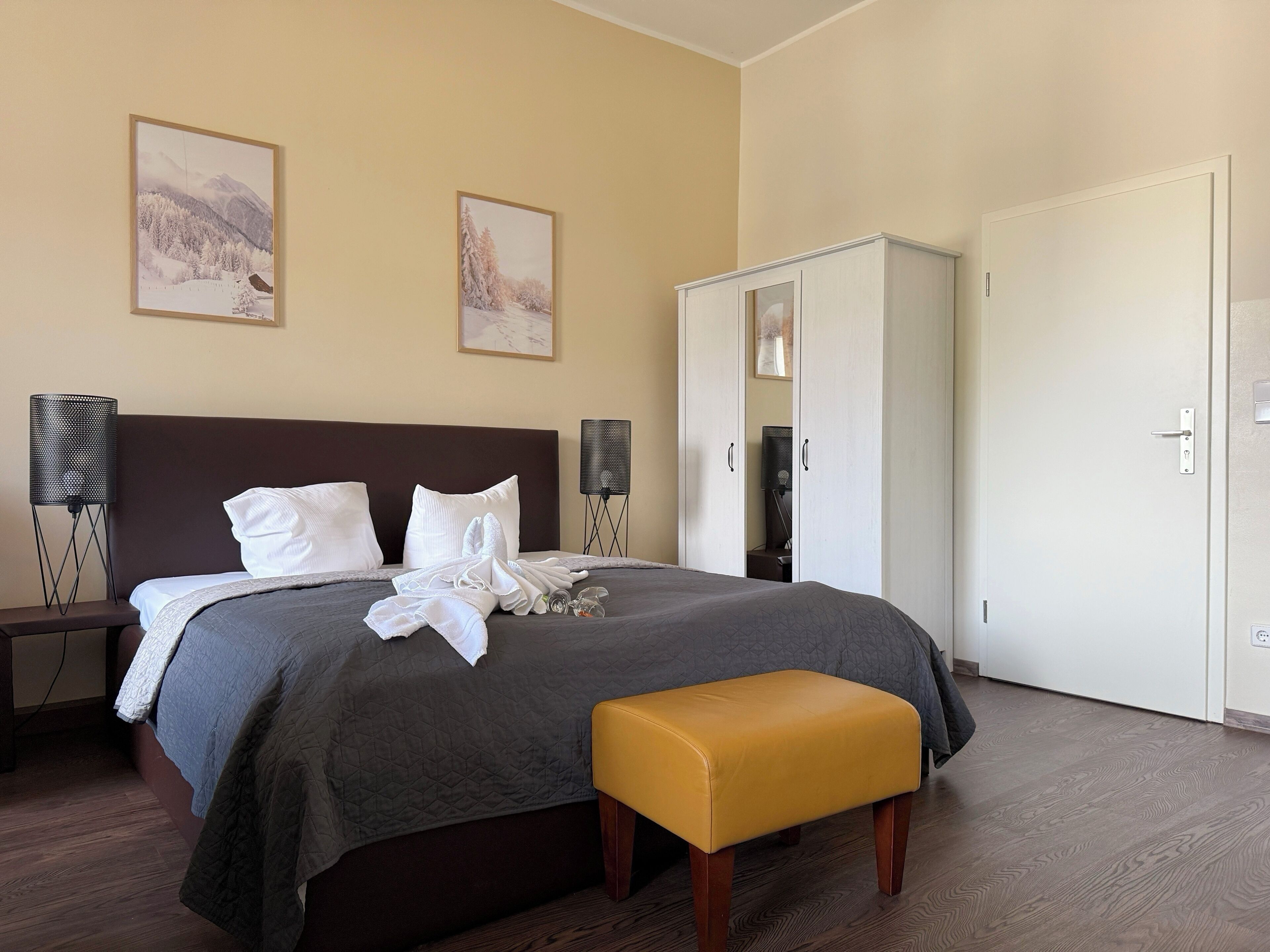 A neatly made bed with pillows, a bedside lamp, and a small table. There is a wardrobe and a bench in the room.