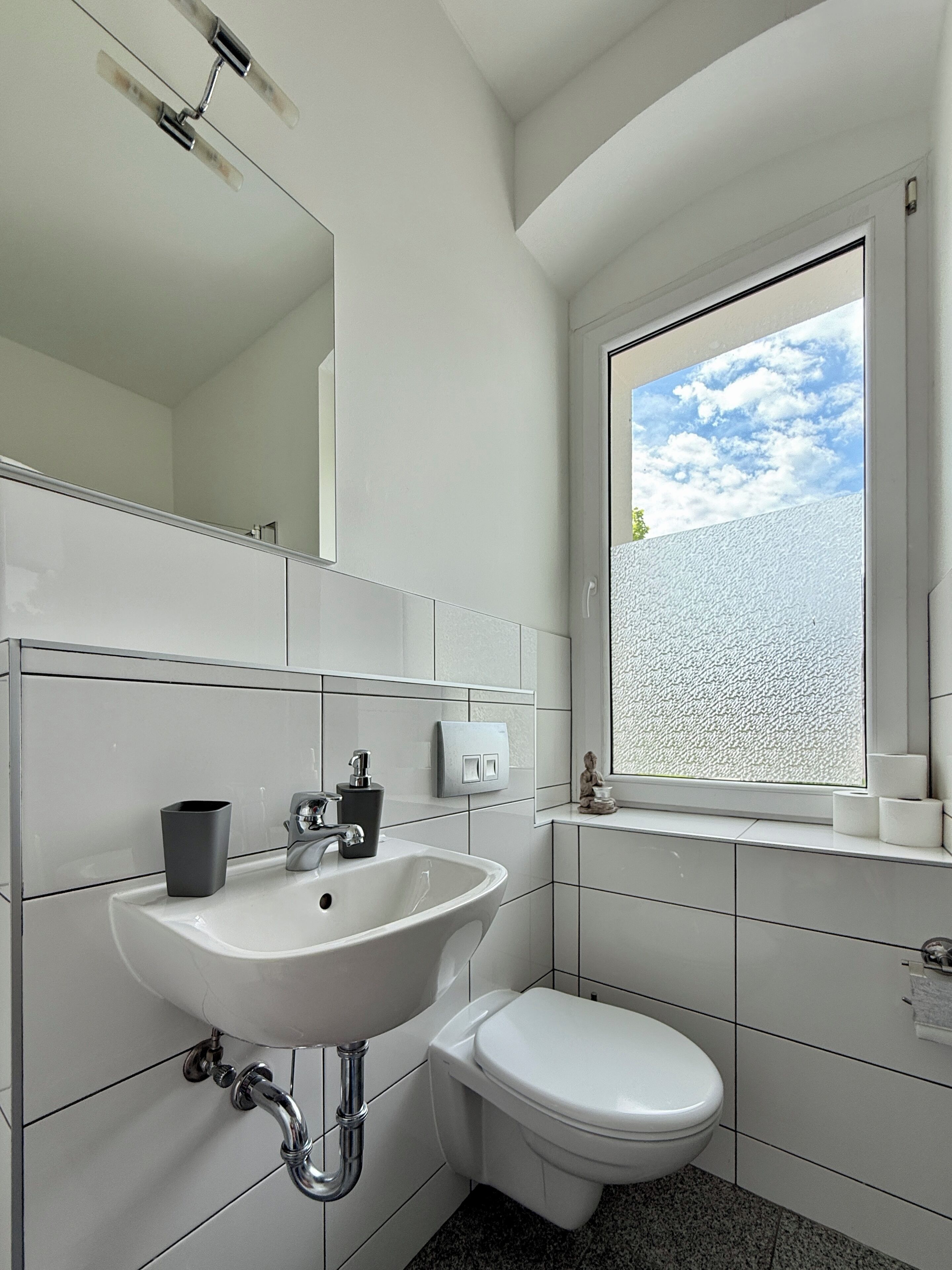 Une salle de bain moderne avec un lavabo blanc, des toilettes et une fenêtre donnant sur le ciel.