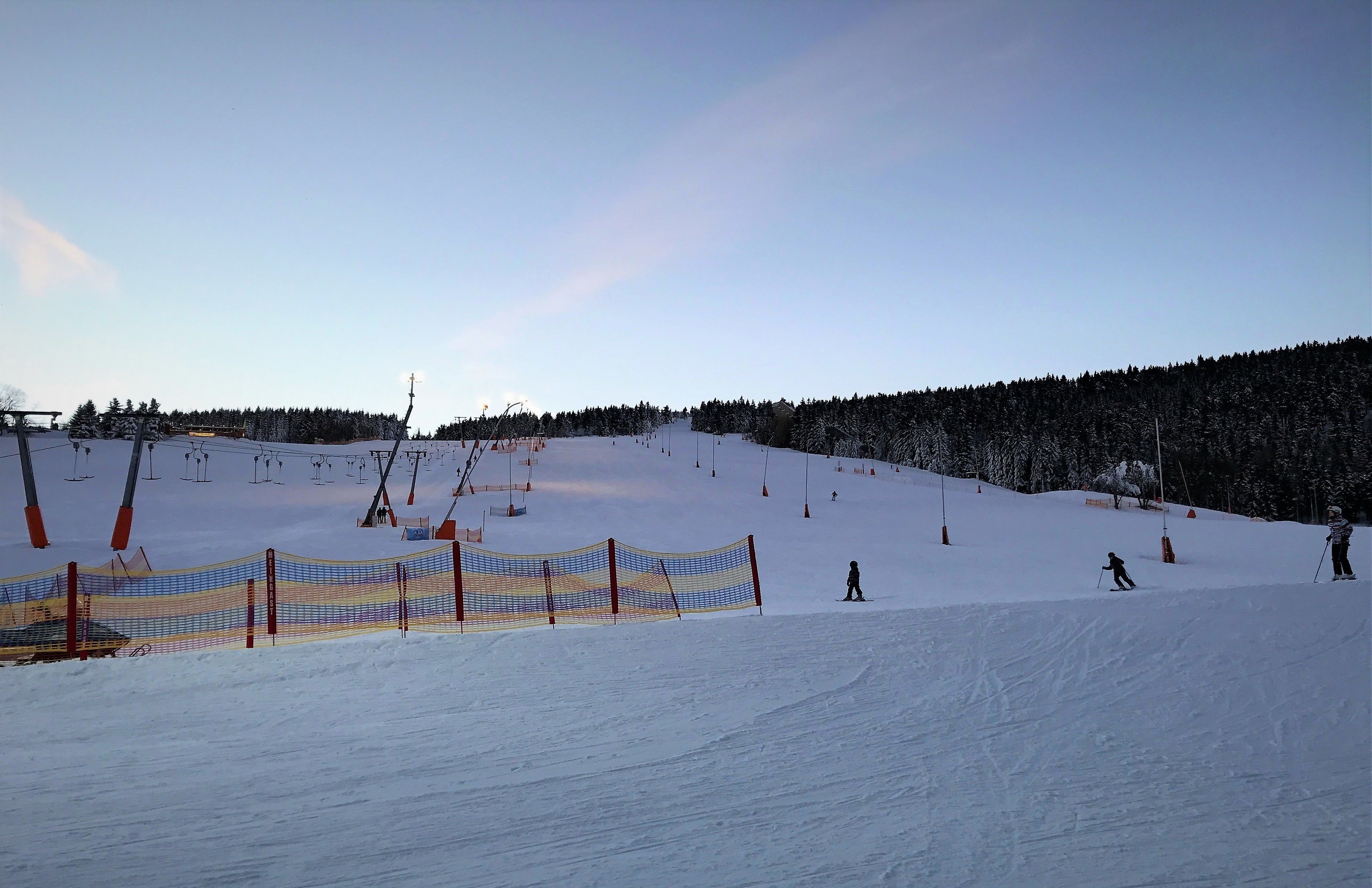 A snowy ski slope with a fence, ski lift, and trees in the background.