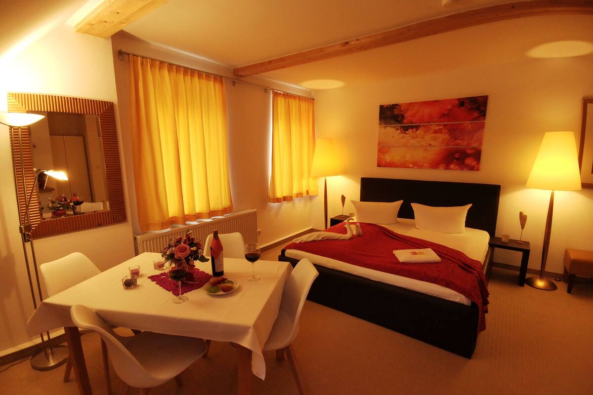 Habitación de hotel con cama, mesitas de noche, escritorio con sillas, mesa de comedor y un amplio ventanal con cortinas.