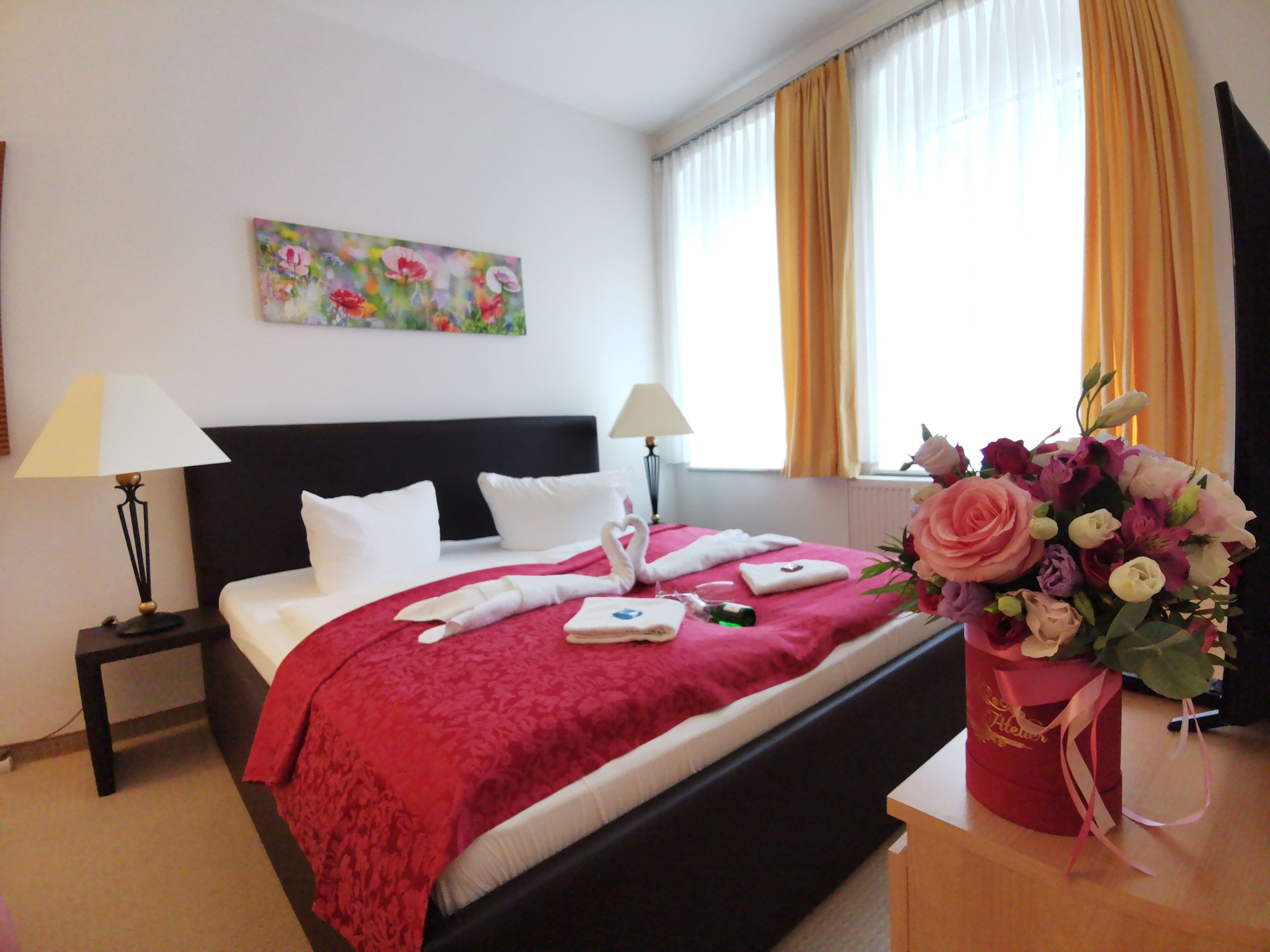 Habitación de hotel con cama, dos mesitas de noche, un cuadro en la pared, ventana con cortinas y un jarrón de flores.