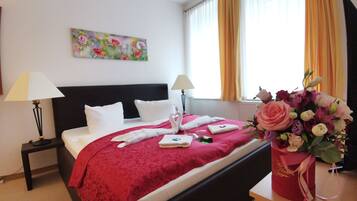 Habitación de hotel con cama, dos mesitas de noche, un cuadro en la pared, ventana con cortinas y un jarrón de flores.