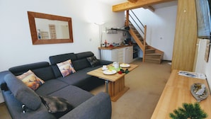 A living room with a sofa, a dining table with plates and a plant, a kitchen area with a sink and cabinets, and a wooden staircase. - Apartments Fichtelberger Blick (Oberwiesenthal)