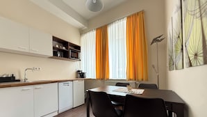 A compact kitchen with white cabinets, a wooden countertop, a sink, and a coffee maker. There is a dining table with two chairs and a window with orange curtains. - Apartments Fichtelberger Blick (Oberwiesenthal)
