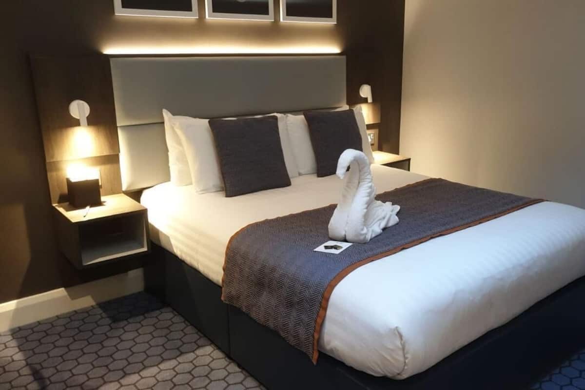 Un lit bien fait, agrémenté d’une serviette blanche en forme de cygne, d’une tête de lit sombre et de deux lampes de chevet.