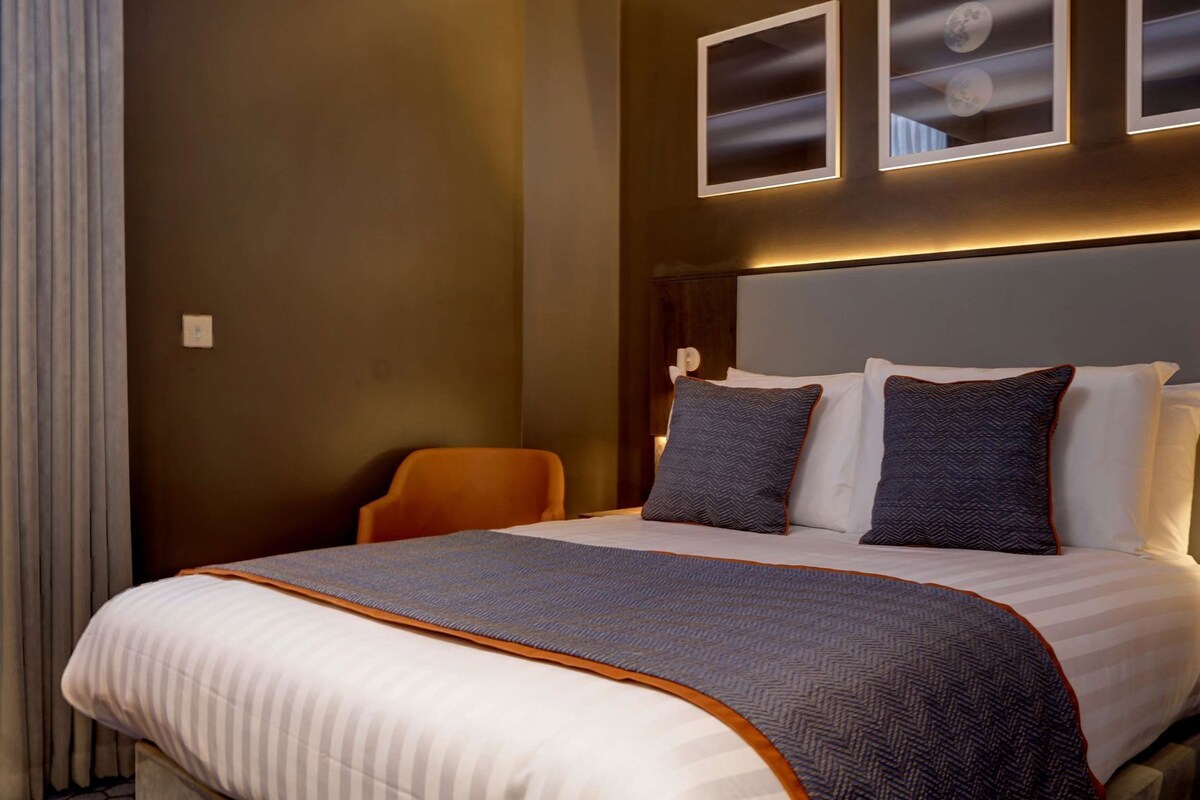 A hotel room with a bed, a chair, and wall-mounted light fixtures.