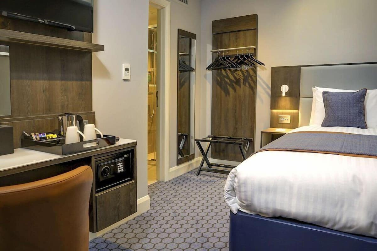 Une chambre d’hôtel comprenant un lit, un bureau, une chaise, une armoire et une petite table avec une lampe.