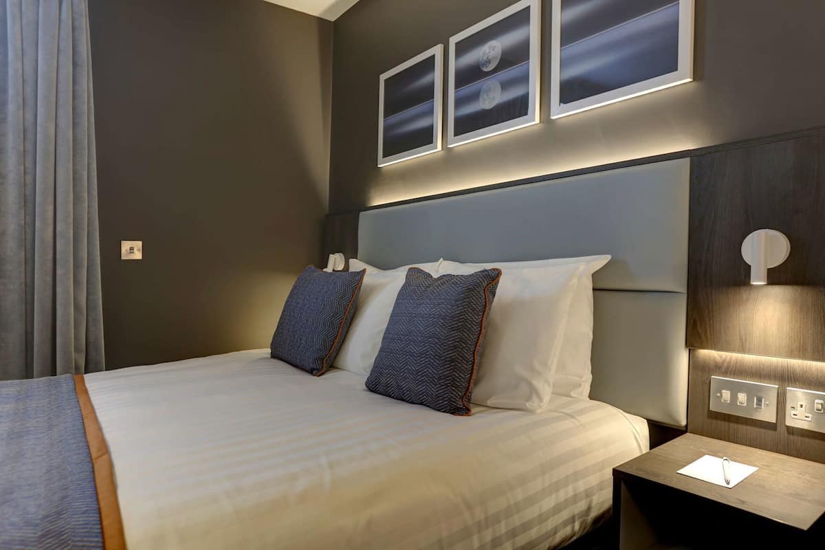 Une chambre d’hôtel avec un lit, deux oreillers, une table de chevet, une lampe et trois tableaux encadrés accrochés au mur.