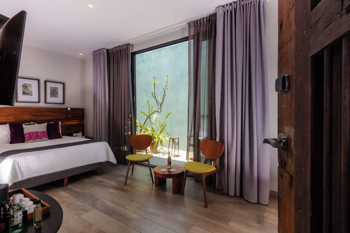 A hotel room with a bed, two chairs, a small table, a potted plant, and a sliding door.
