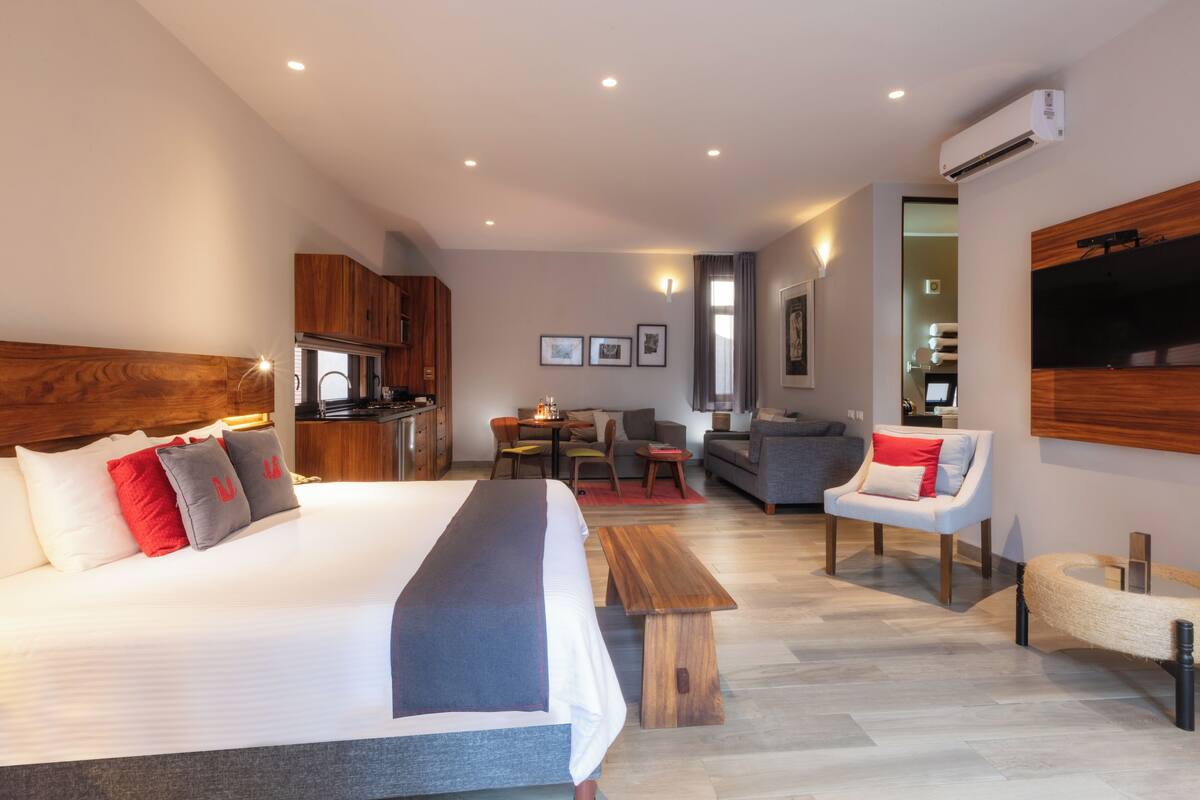 A modern hotel room with a large bed, a wooden headboard, a dining area, a sofa, and a TV mounted on the wall.