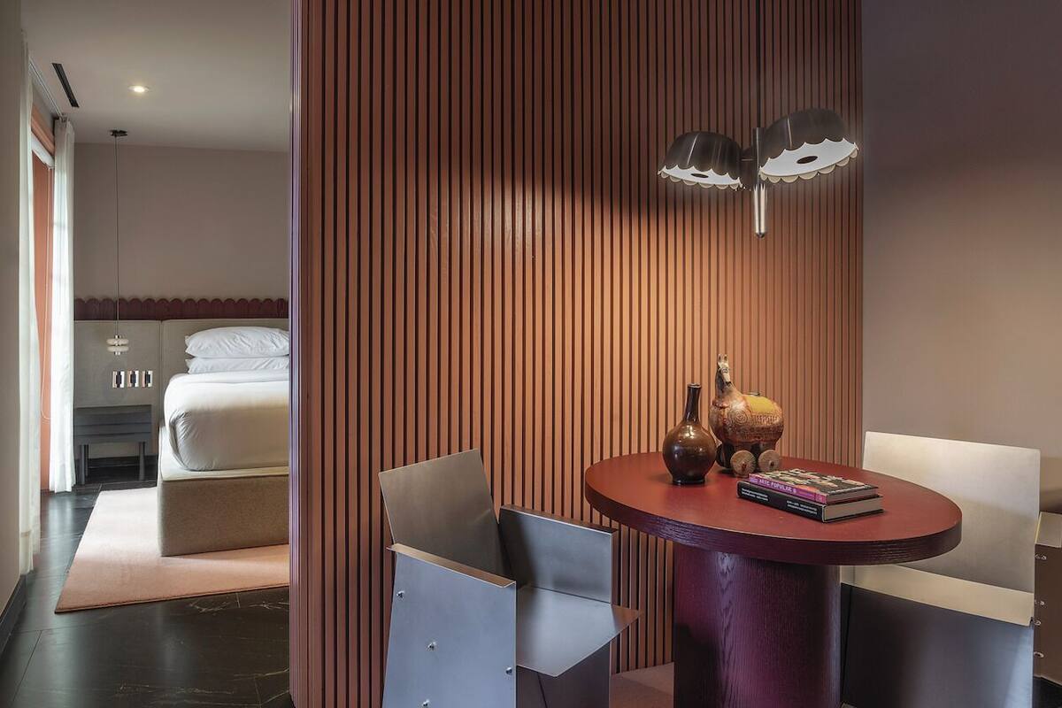 Une chambre d’hôtel moderne avec un lit, une table ronde sur laquelle sont posés des livres, un vase et une lampe décorative.
