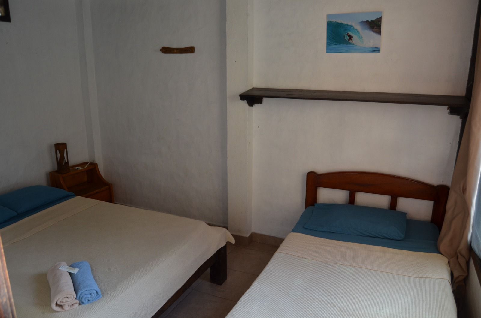 Habitación con dos camas, cabecera de madera, mesita de noche pequeña, estante en la pared y un cuadro de un surfista.