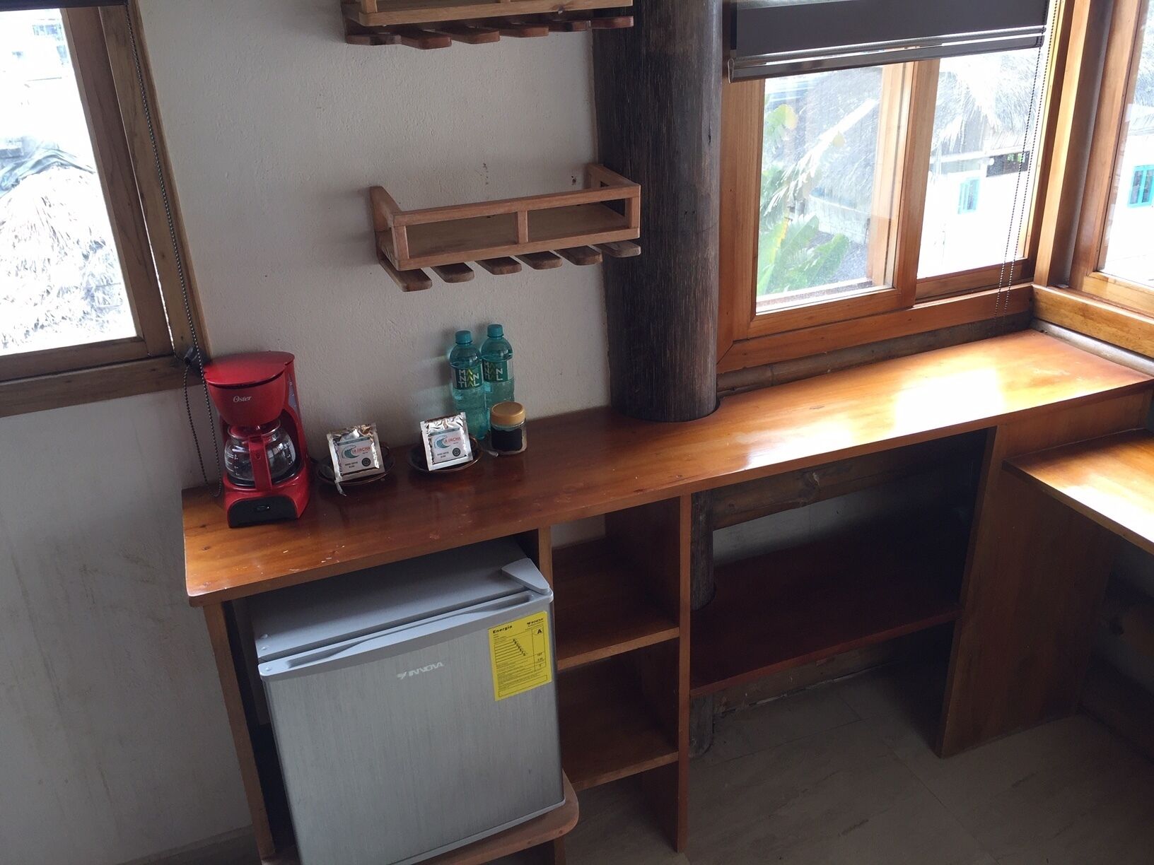 Una kitchenette compacta con refrigerador, cafetera y una repisa con agua embotellada y un recipiente.