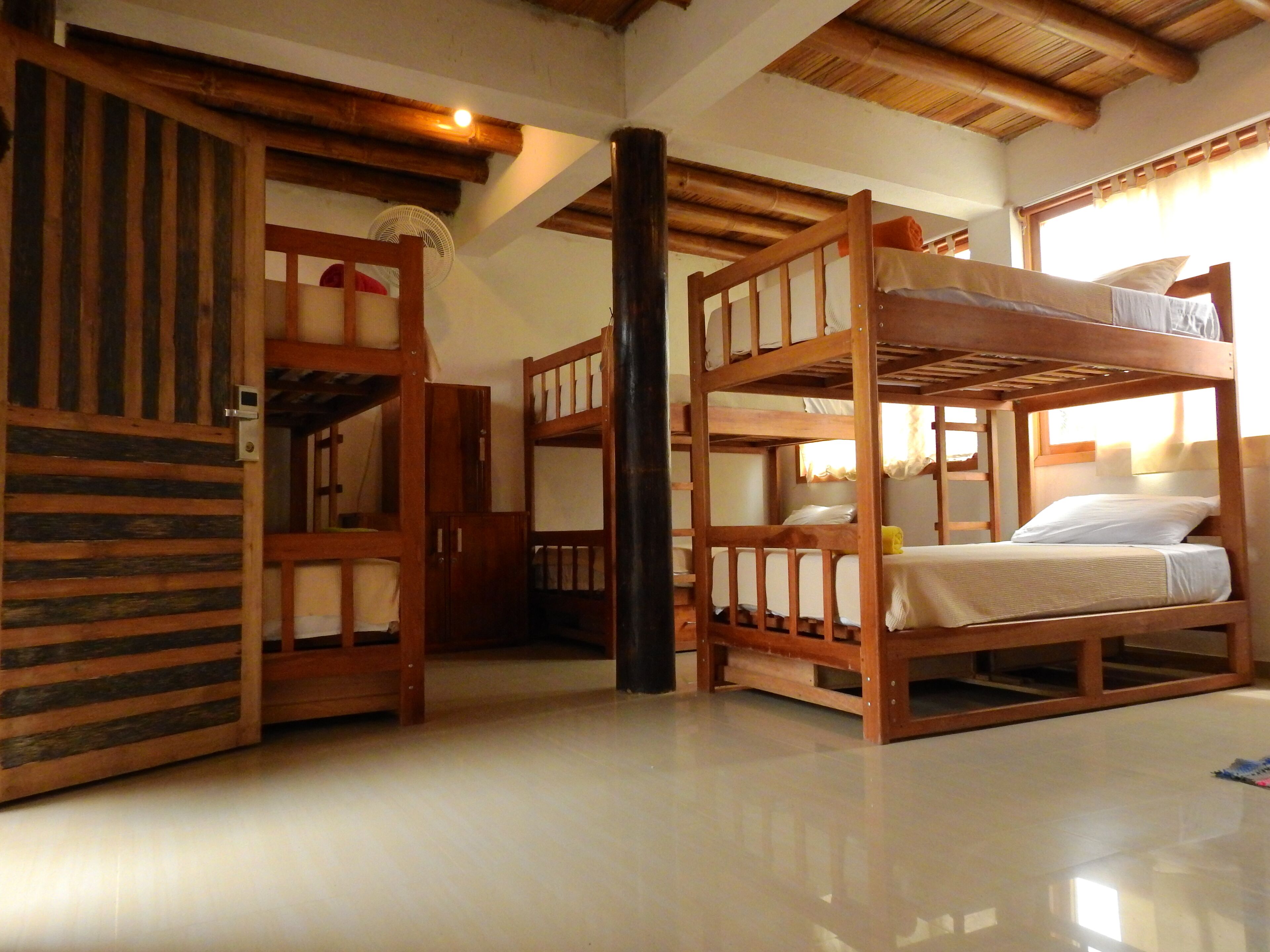 Quarto com beliches, vigas de madeira, um ventilador e uma janela com cortinas.