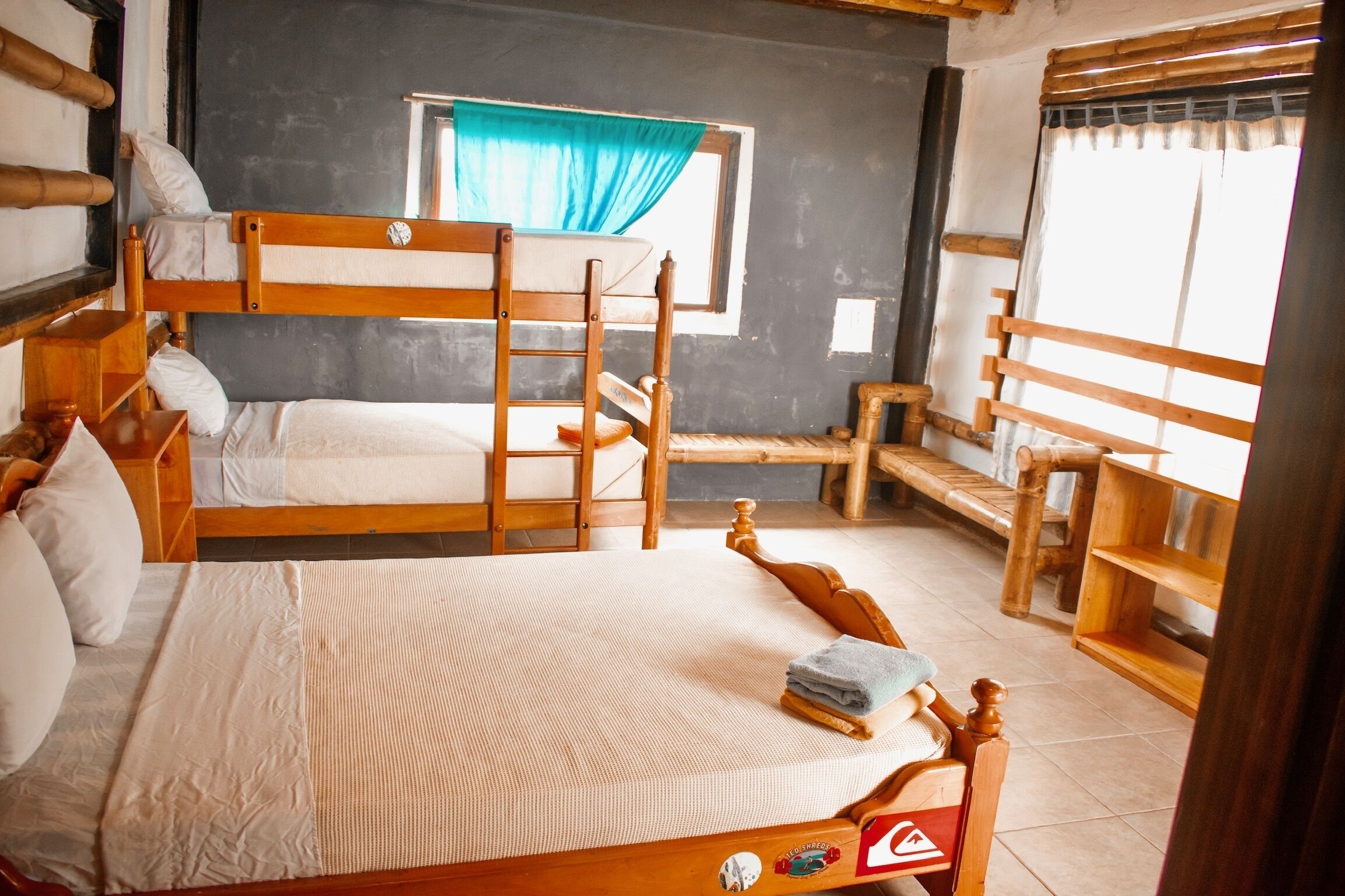 A dormitory room with bunk beds, a window with blue curtains, and a wooden ladder.