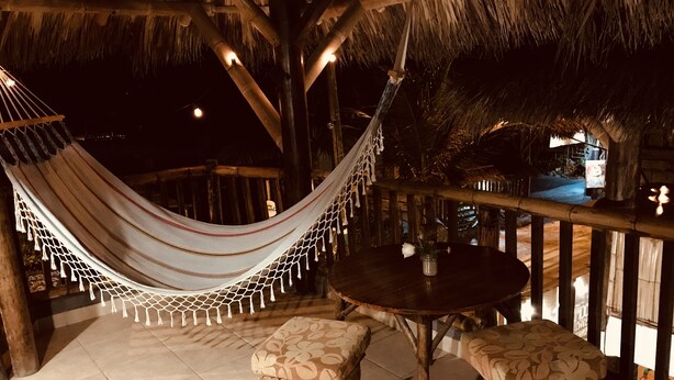 A hammock, two wooden stools, and a round table on a tiled patio with a thatched roof. - La Facha Hostel Restaurant Surf (Muisne)