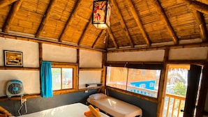 A room with a thatched roof, wooden beams, a bed with white linens, a bedside table with a lamp, and a window with a view of buildings outside. - La Facha Hostel Restaurant Surf (Muisne)
