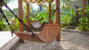 Una hamaca de madera columpiándose bajo una pérgola de madera, rodeada de abundante follaje y un camino empedrado.