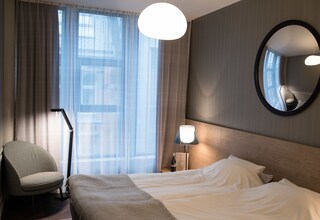 A hotel room with a bed, a chair, a lamp, a mirror, and a window with curtains.