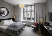 A hotel room with a bed, a desk, a chair, a large window with curtains, a round mirror, and a hanging light fixture.