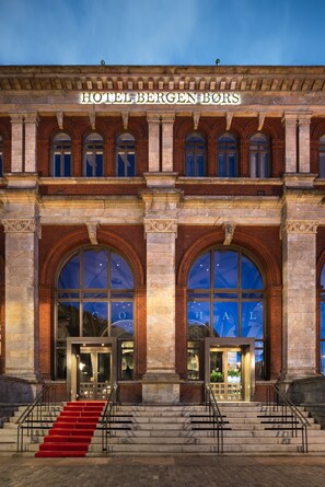 A historic building with the sign "HOTEL BERGENBORS" at the top. It features large arched windows and a grand entrance with a red carpet leading up to the doors.