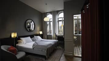 A hotel room with two beds, a sitting area with a chair, a desk, and a bathroom visible through an open door.