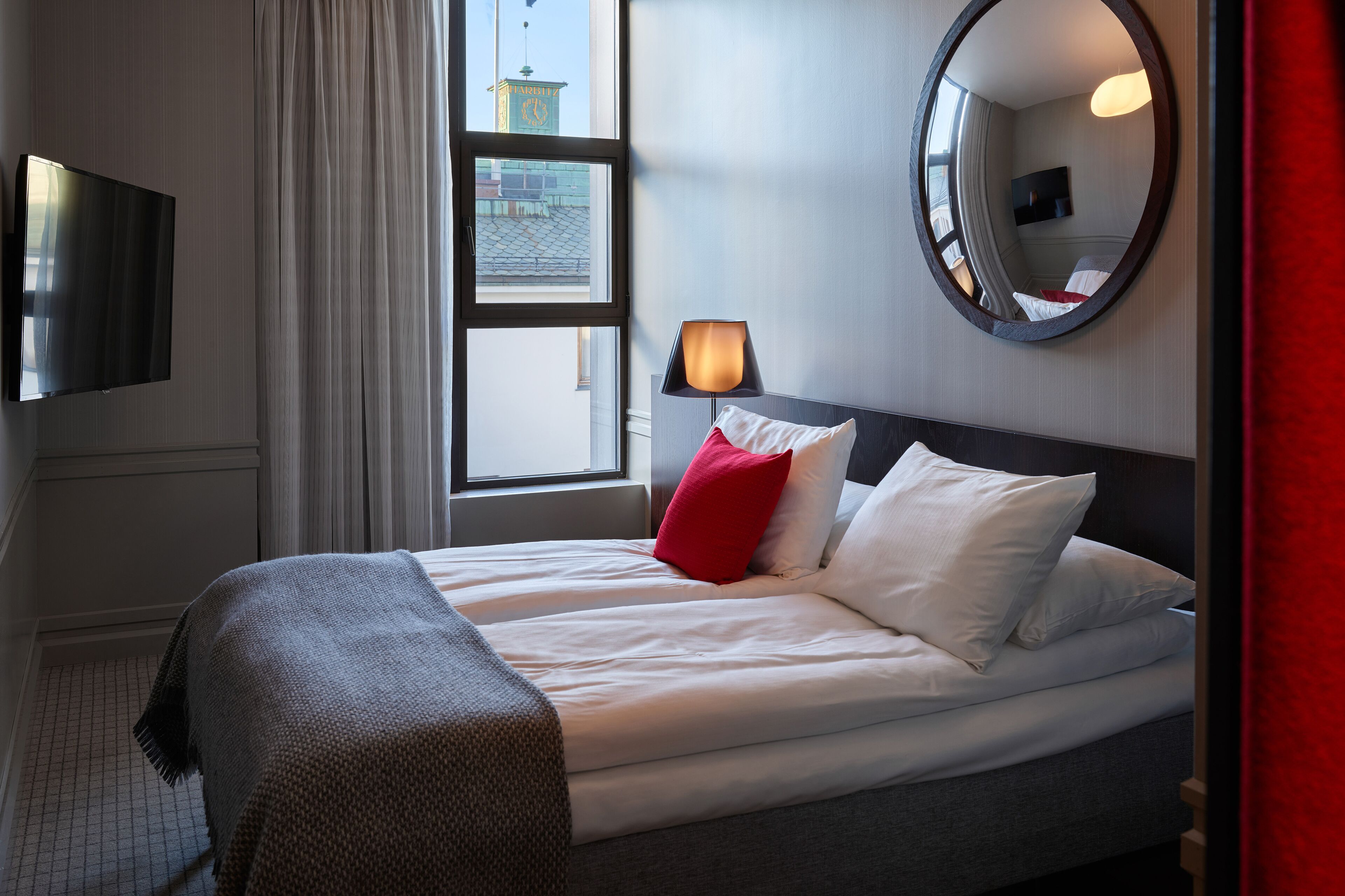 A hotel room with a bed, a TV, a window, and a mirror.