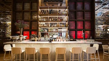 Een bar met een marmeren aanrecht, meerdere flessenplanken en verschillende witte barstoelen.