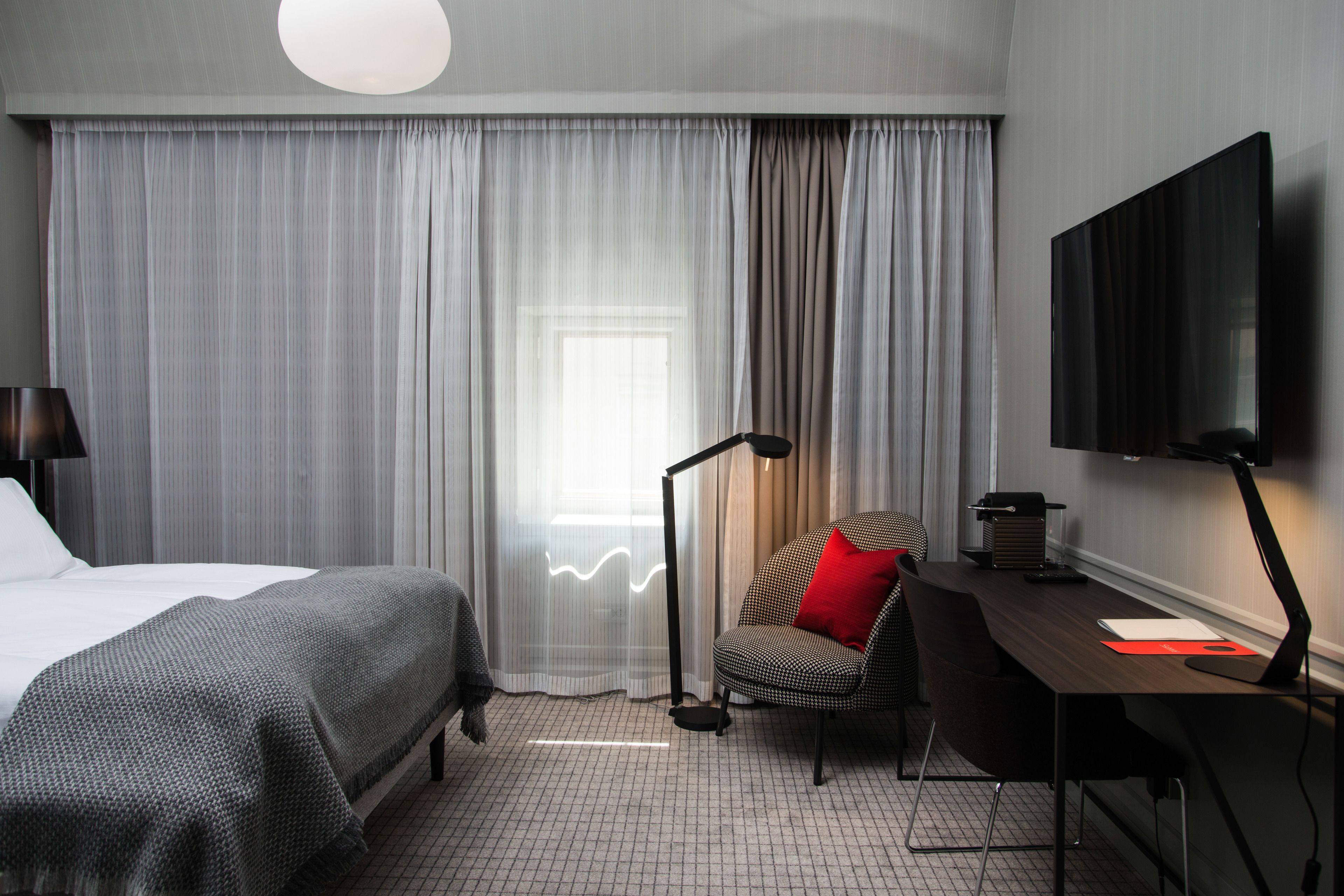 Habitación de hotel con cama, escritorio, silla, televisor y ventana con cortinas.
