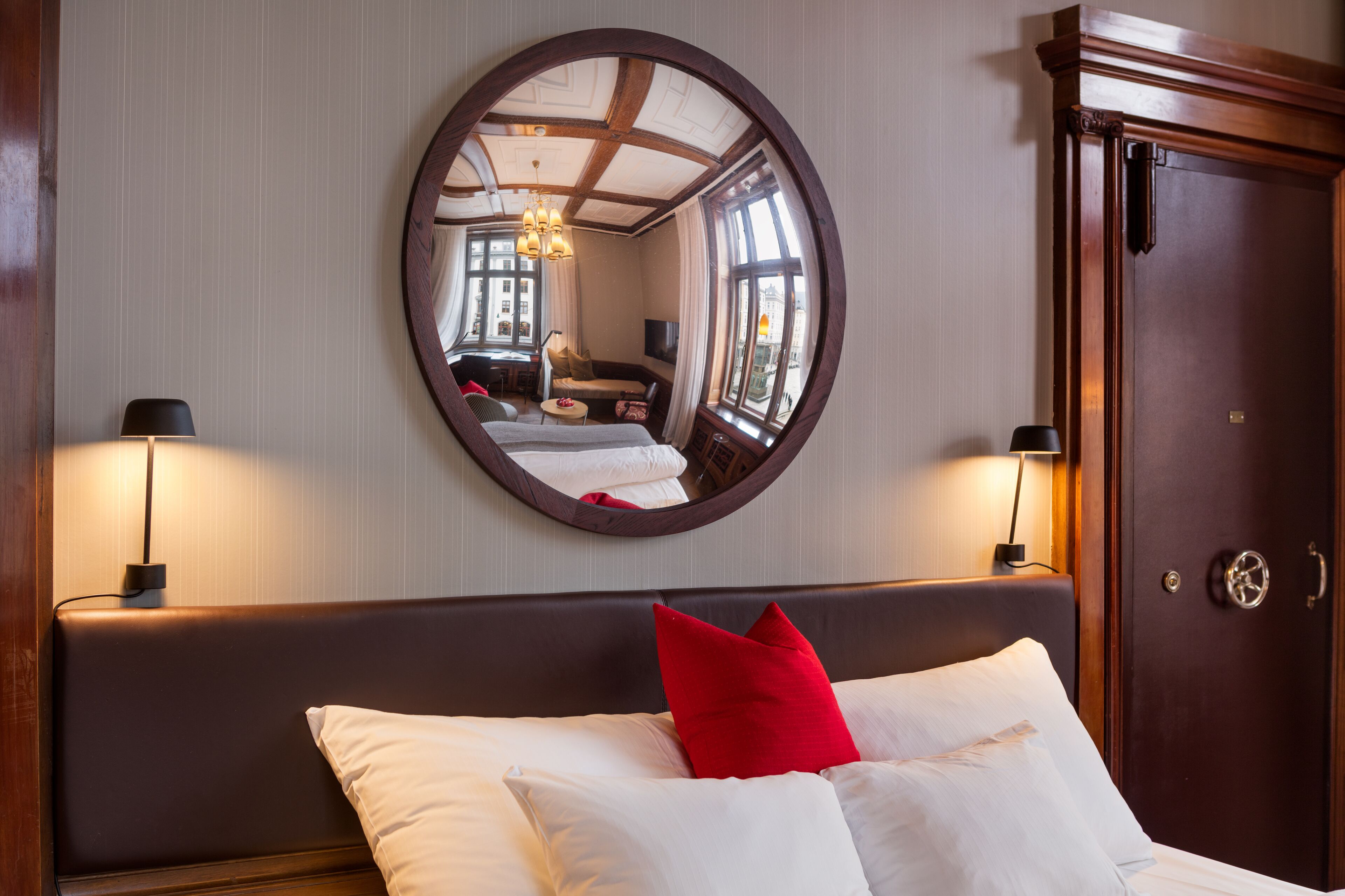 Une chambre d’hôtel avec un grand miroir rond, un lit avec des oreillers blancs et rouges, une tête de lit en bois et une armoire en bois.