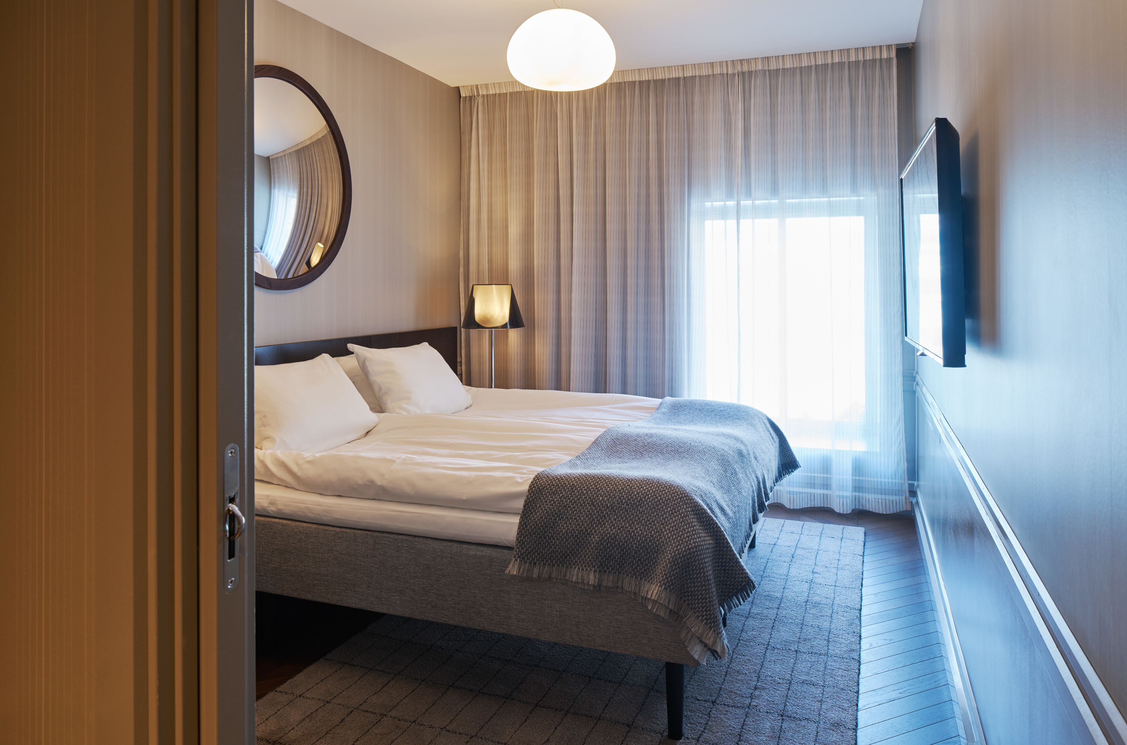Habitación de hotel con cama, lámpara de noche, televisor montado en la pared y ventana con cortinas.
