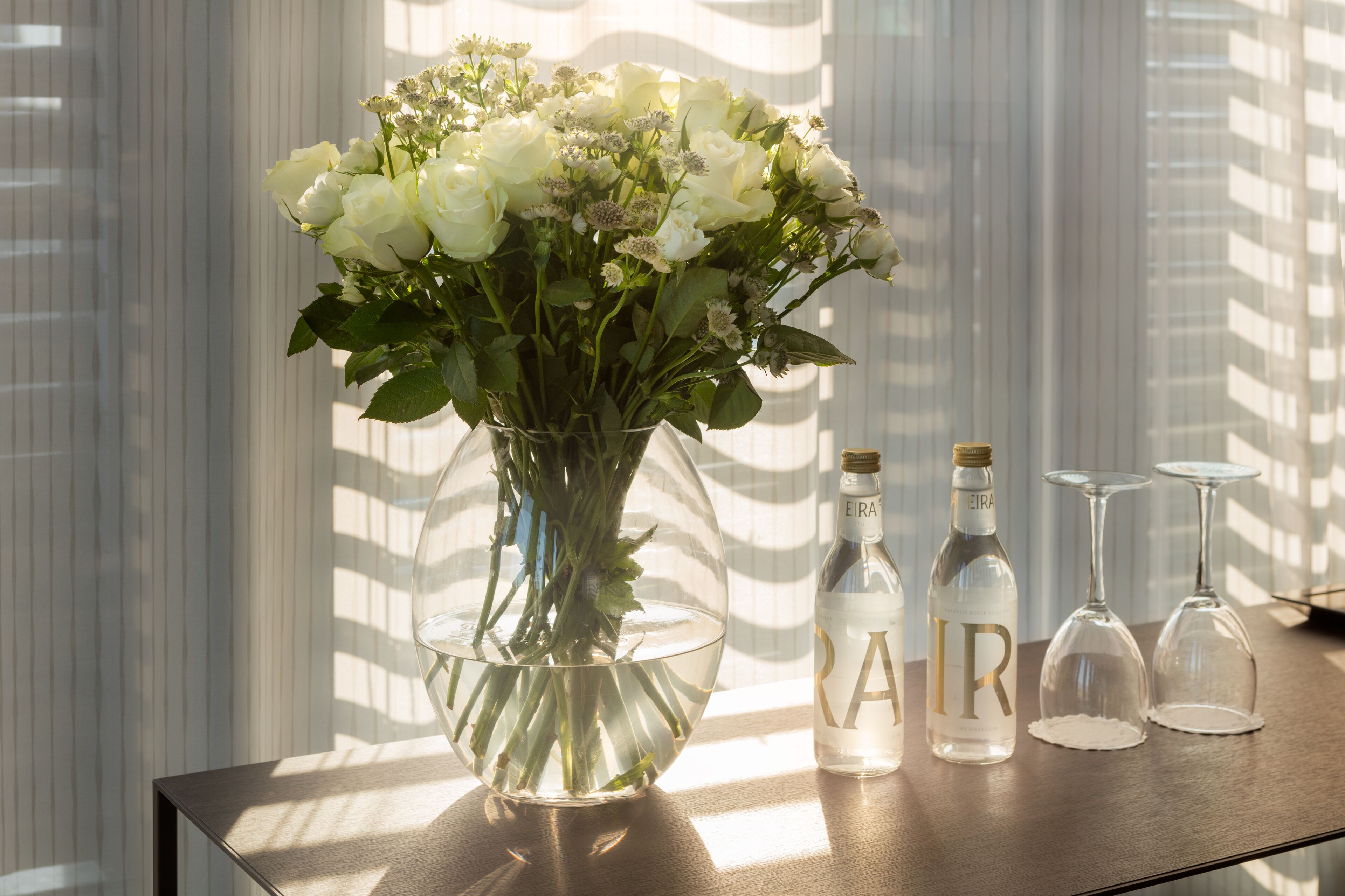 Un jarrón de cristal transparente con flores blancas sobre una mesa de madera, junto a una botella y dos copas de vino.