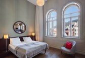 A hotel room with a large bed, two bedside lamps, a round mirror, and two large windows with a view of buildings.