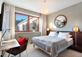 A hotel room with a bed, bedside lamps, a desk with a chair, a window with curtains, and a mirror on the wall.