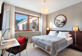 A hotel room with a bed, bedside lamps, a desk with a chair, a window with curtains, and a mirror on the wall.