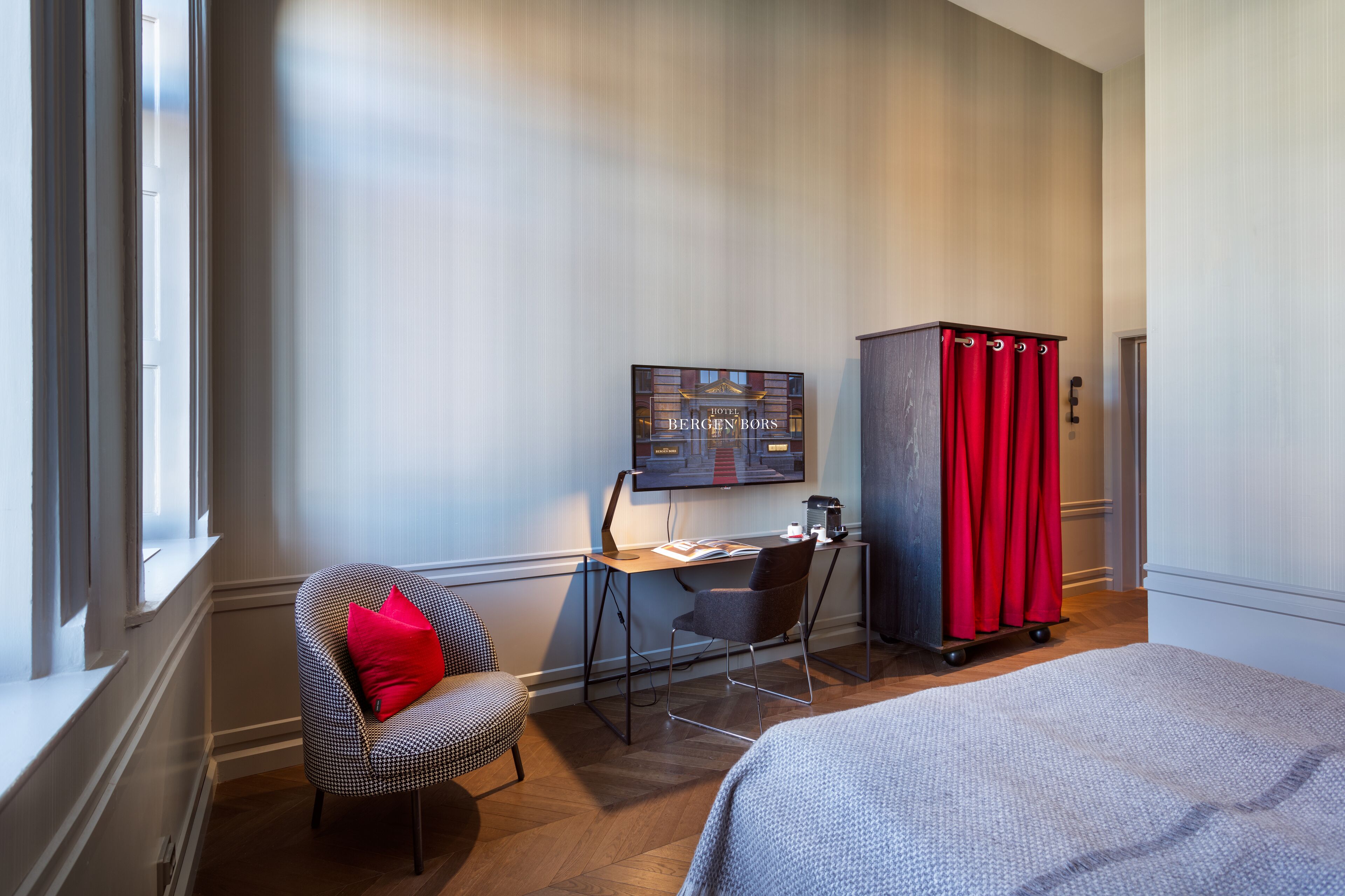 A hotel room with a desk, a chair with a red pillow, a television, and a red cabinet.