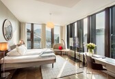 A modern hotel room with a large bed, a desk, a chair, a lamp, and a view of the city through large windows.