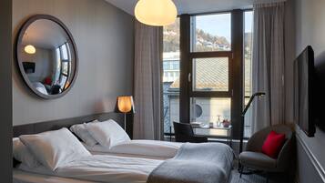 Ein Hotelzimmer mit einem großen Bett, einem Sessel, einem Tisch, einem Spiegel und einem Fenster mit Blick auf Gebäude.