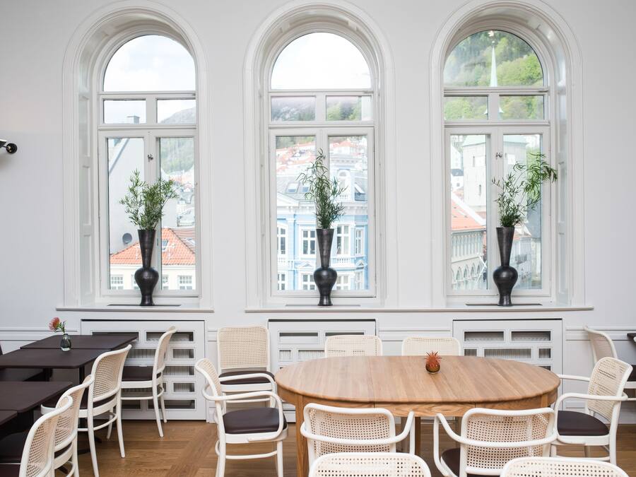Un comedor con una mesa redonda de madera rodeada de sillas de mimbre blanco, ventanas arqueadas amplias con plantas en macetas y vistas a edificios.