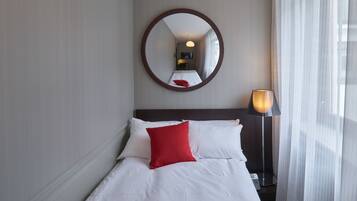 A neatly made bed with white linens and a red pillow, a round mirror on the wall, and a lamp on a bedside table.