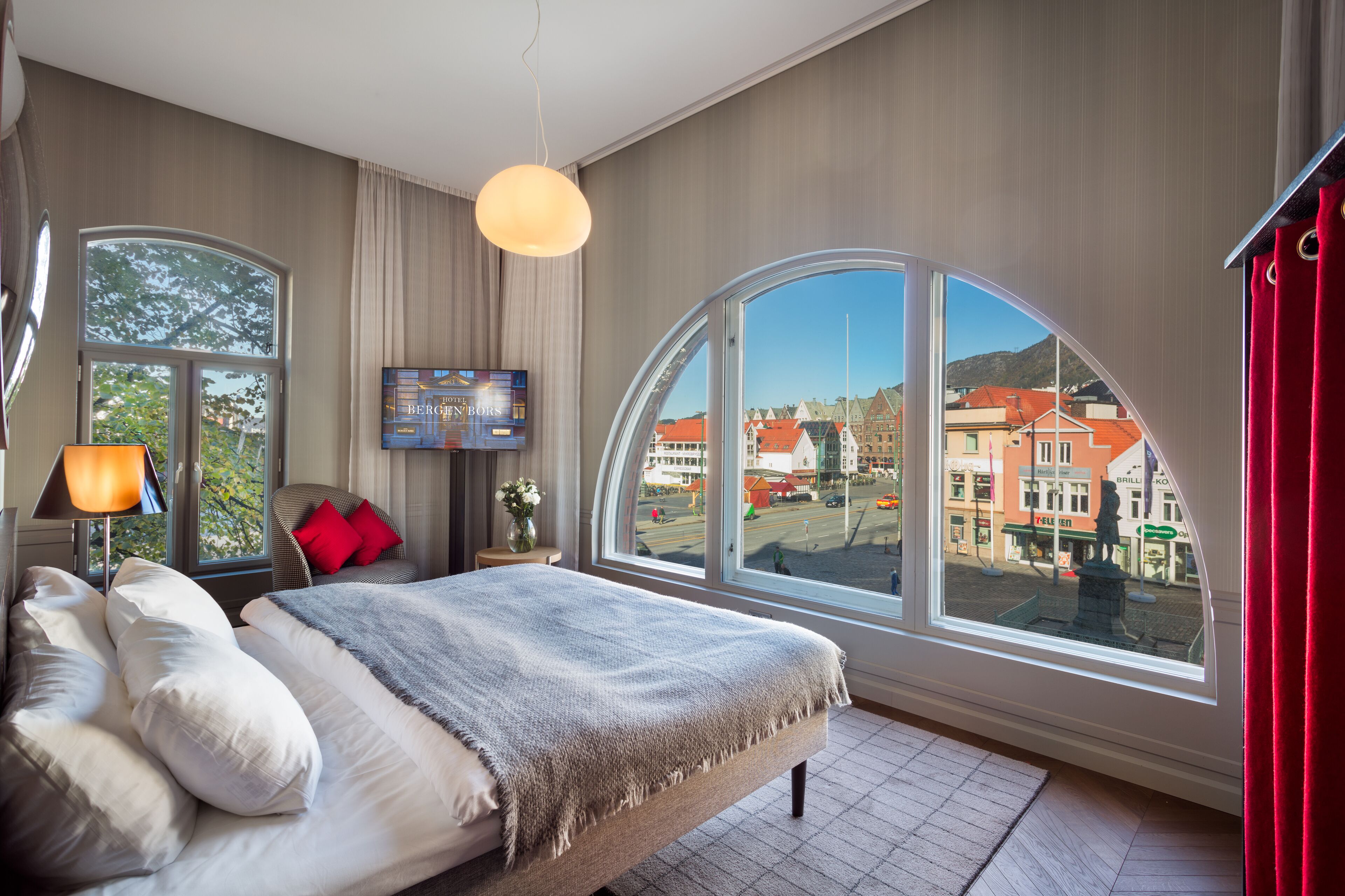 A modern hotel room with a large bed, a TV, and a view of a town through a large window.